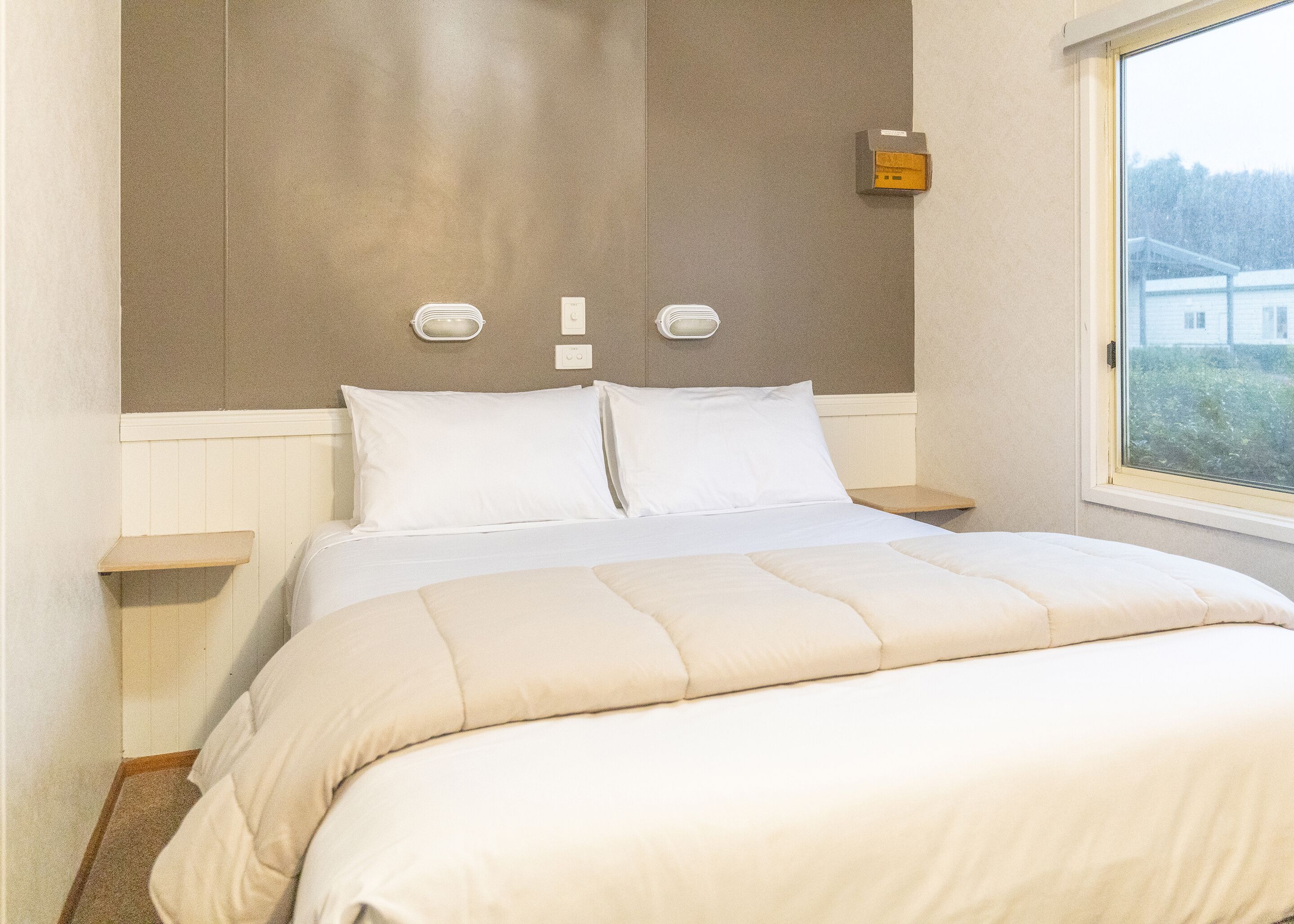 Una cama bien hecha con edredón color beige y almohadas, una mesita de noche con lámpara, y una ventana con vistas a edificios.