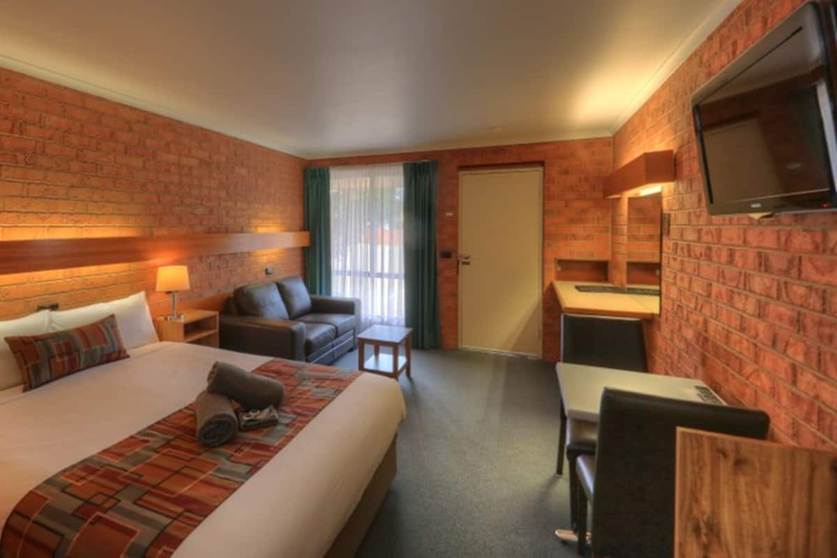 A hotel room with a brick wall, a bed with a patterned bedspread, a sofa, a small table, a TV mounted on the wall, and a door with curtains.