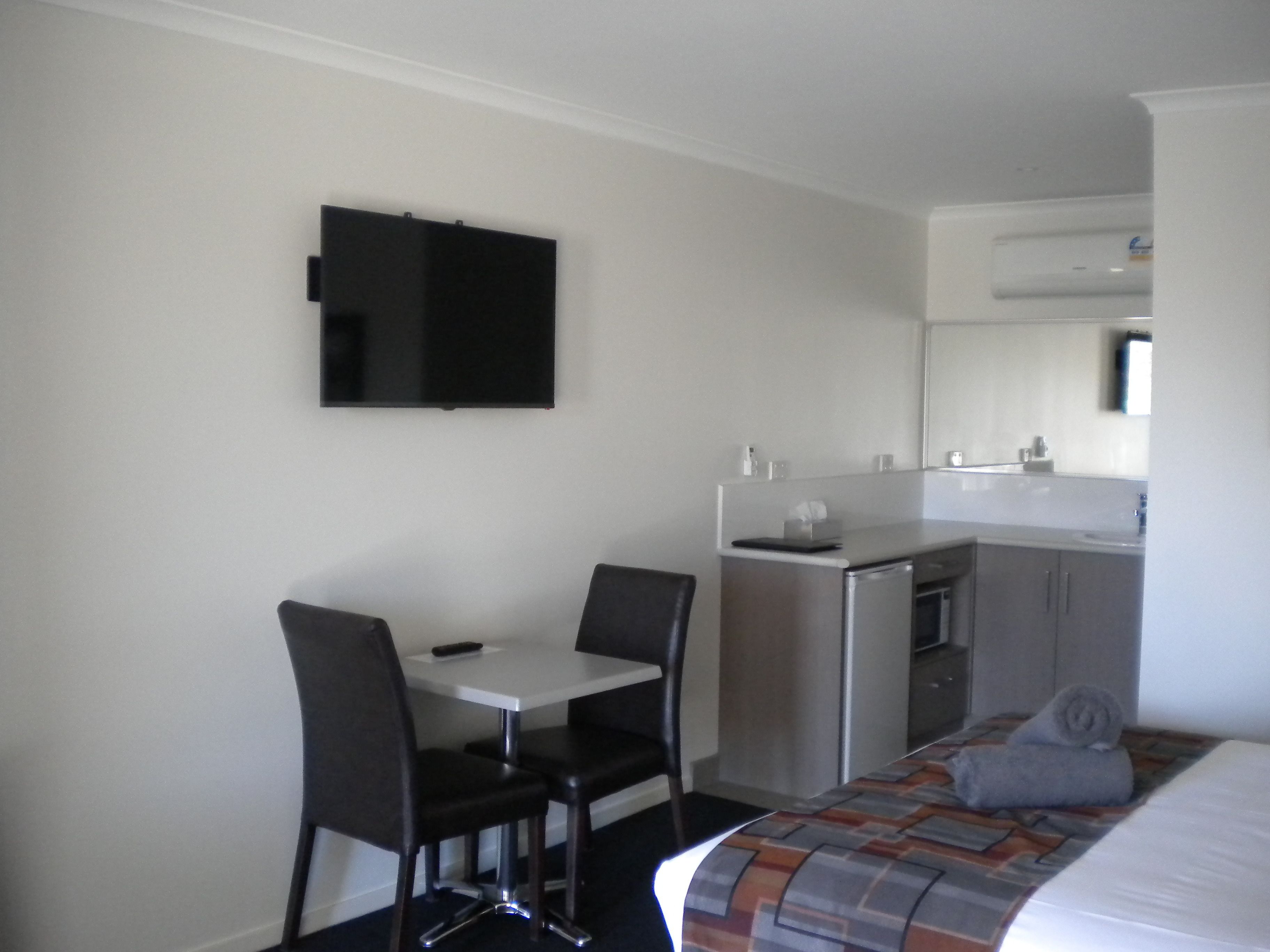 A hotel room with a bed, a desk with two chairs, a small kitchenette, and a flat-screen TV mounted on the wall.