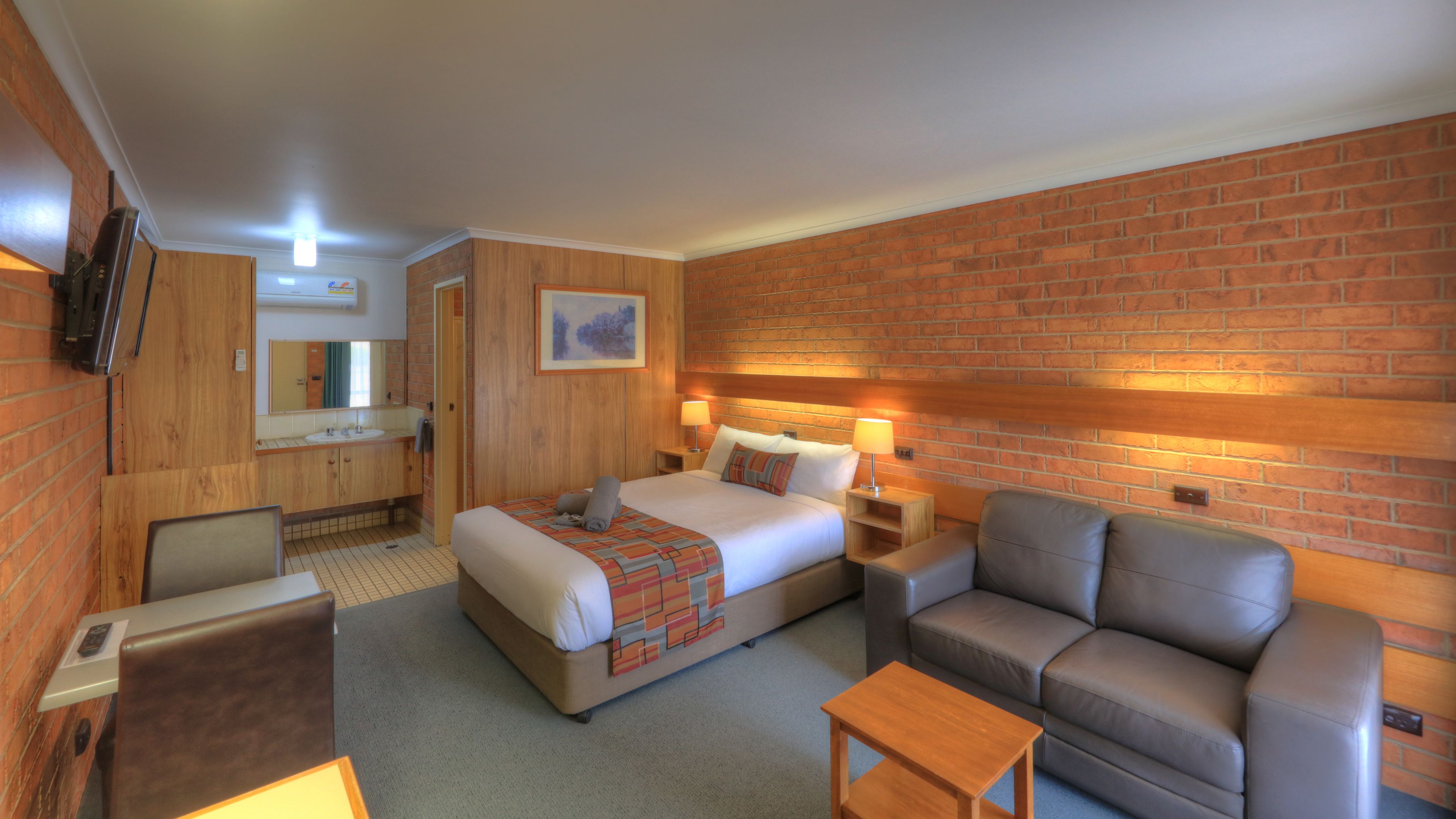 Quarto de hotel com cama, sofá, uma pequena mesa, televisão, banheiro com pia e espelho, e uma parede de tijolos.