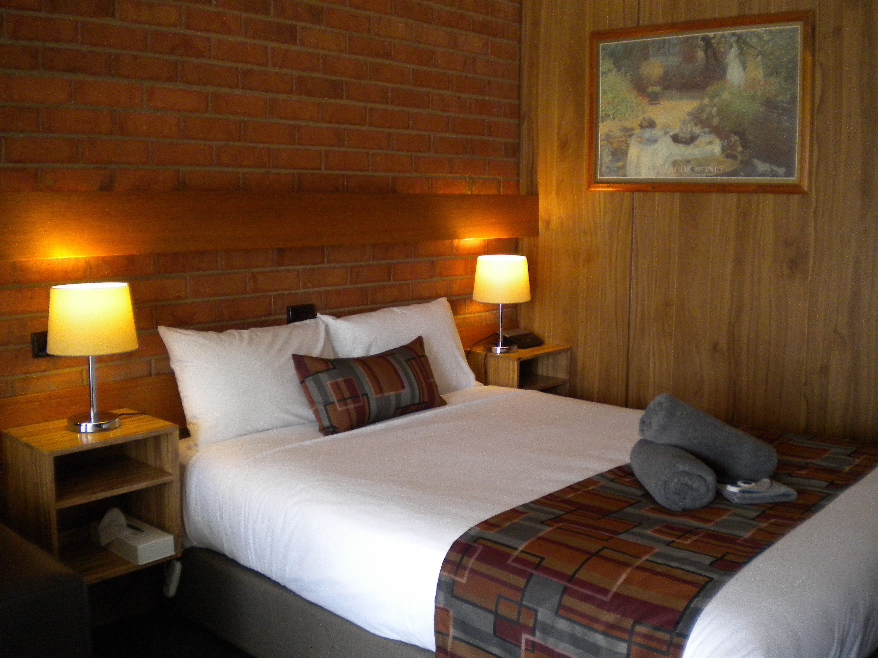Habitación de hotel con pared de ladrillo, dos lámparas de noche, cama con ropa blanca y una manta de cuadros, además de un cuadro en la pared.