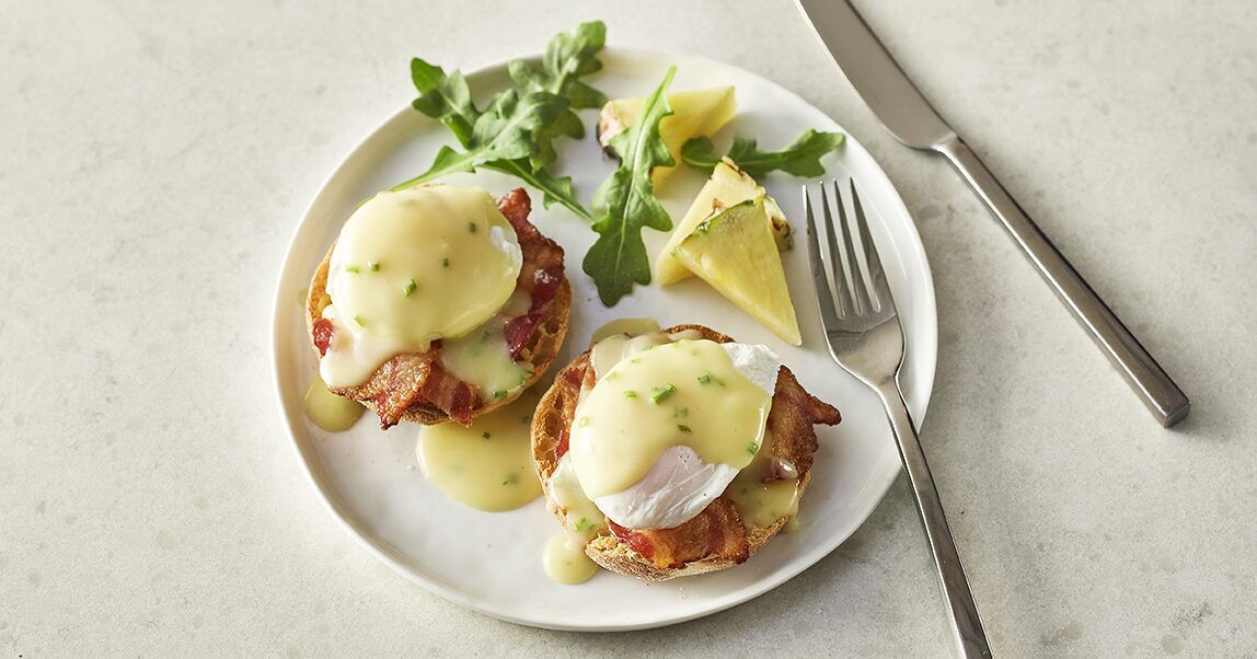 A plate with two eggs benedict sandwiches, garnished with chives, accompanied by a fork and knife on a white plate.