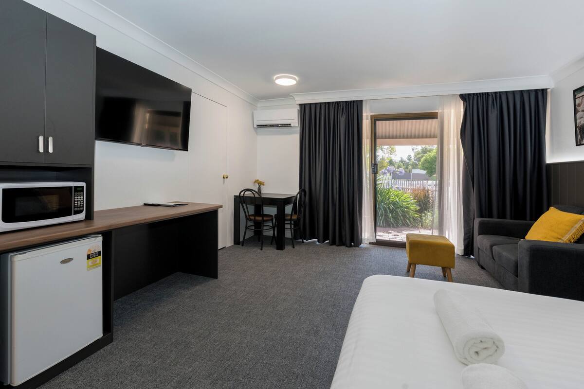 A hotel room with a bed, a desk with a microwave, a small refrigerator, a TV, a dining table with chairs, and a sofa with a yellow pillow.