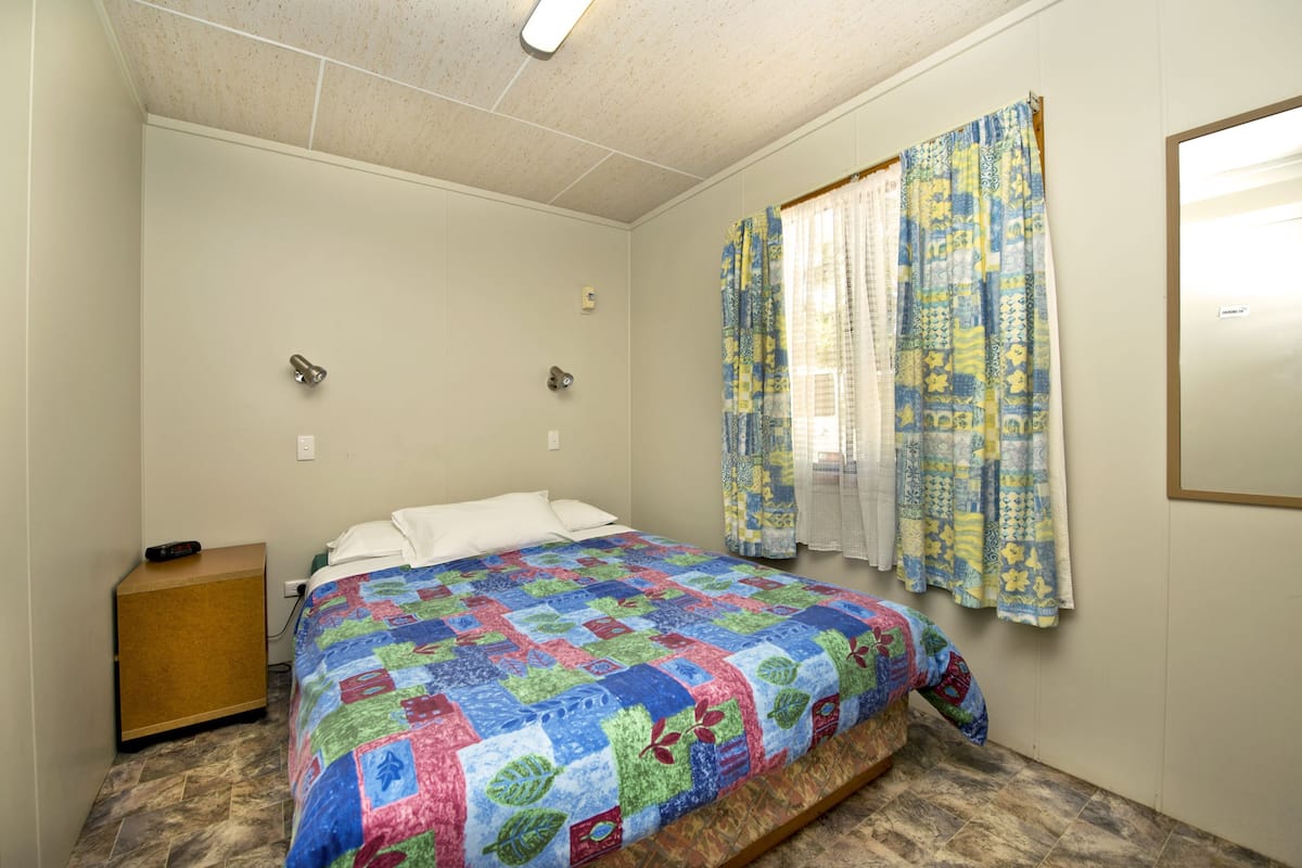 A bedroom with a bed, a nightstand, a window with patterned curtains, and a light fixture on the ceiling.