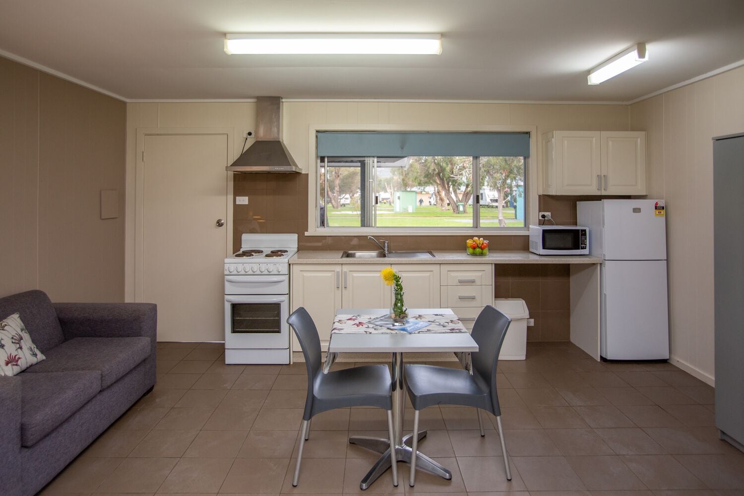 A compact kitchen with a stove, microwave, and refrigerator. There is a dining table with two chairs and a vase with flowers. A sofa is in the corner, and a window provides a view of the outdoors.