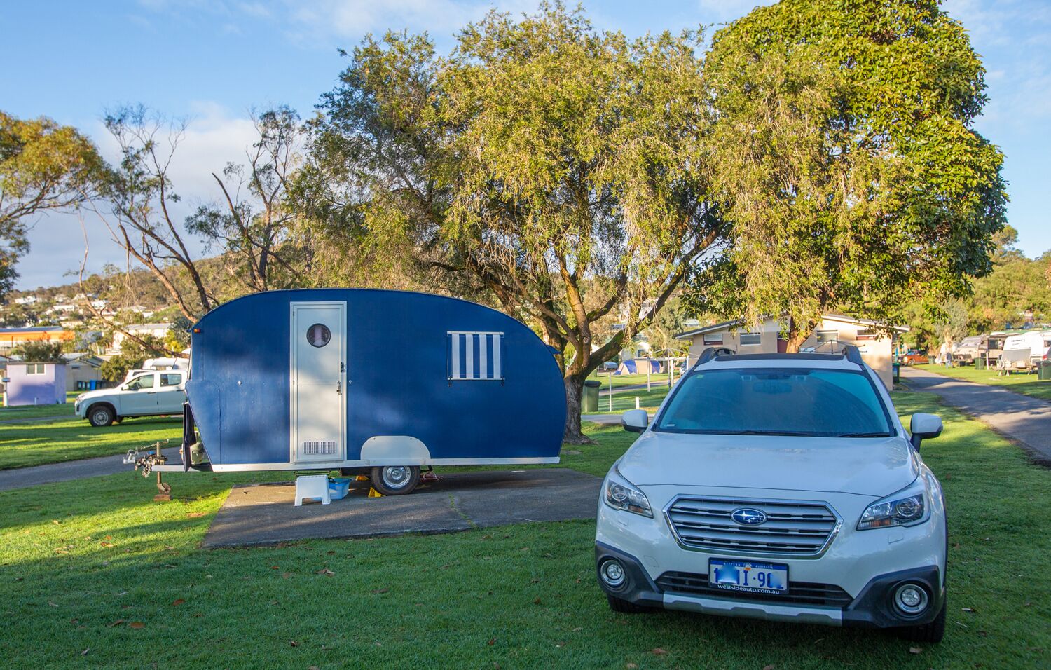 A white SUV parked next to a blue recreational vehicle in a grassy area with trees and houses in the background.
