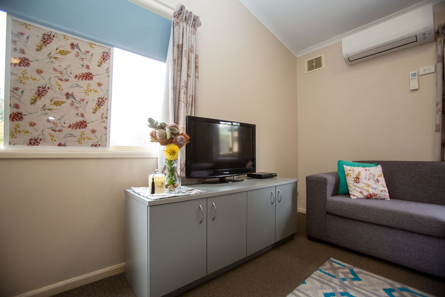 A living room with a grey sofa, a flat-screen TV on a grey cabinet, a window with floral curtains, and an air conditioner on the wall.