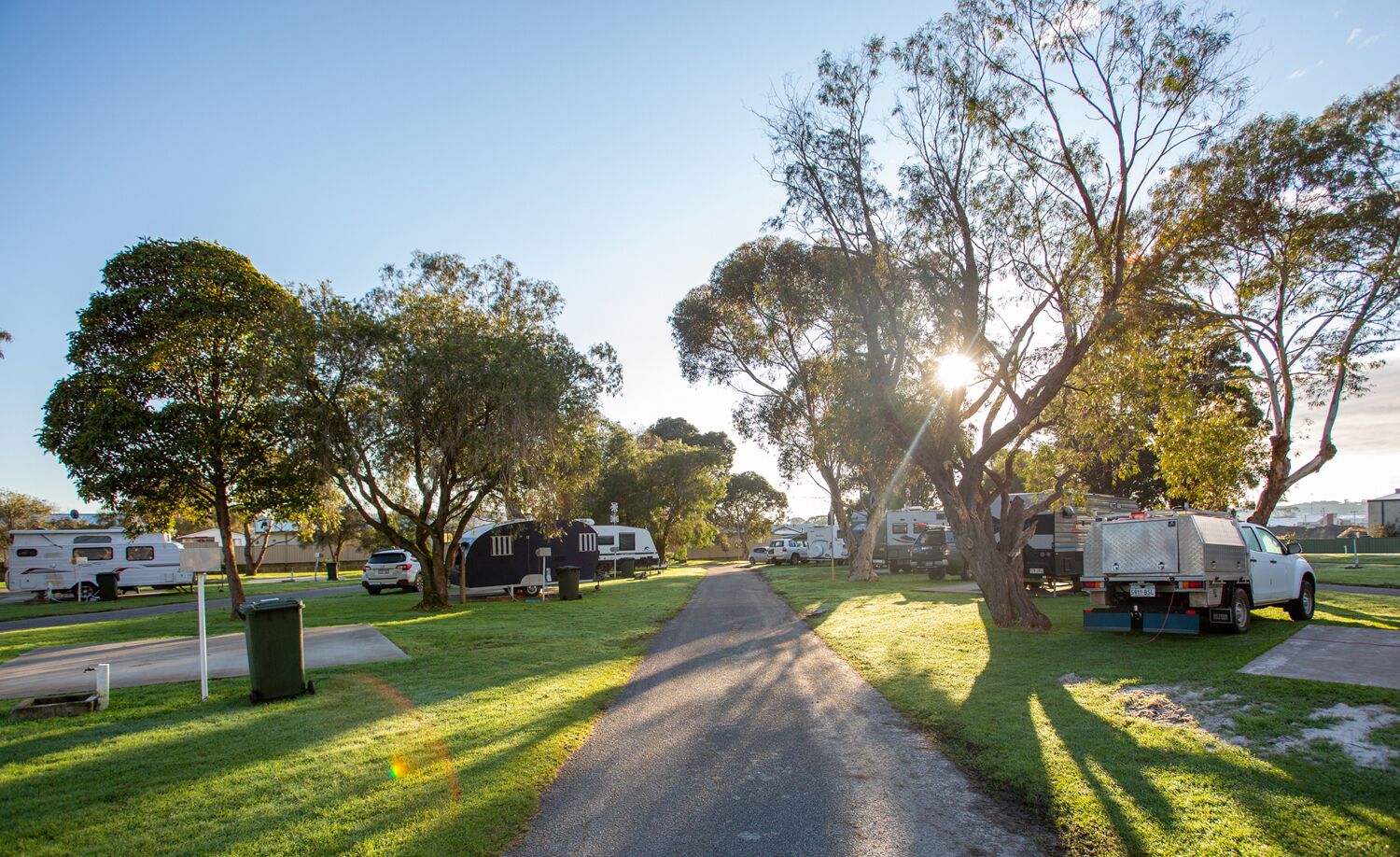 Un terrain de camping où des camping-cars sont stationnés sur des pelouses, avec un chemin pavé et des arbres qui offrent de l’ombre.