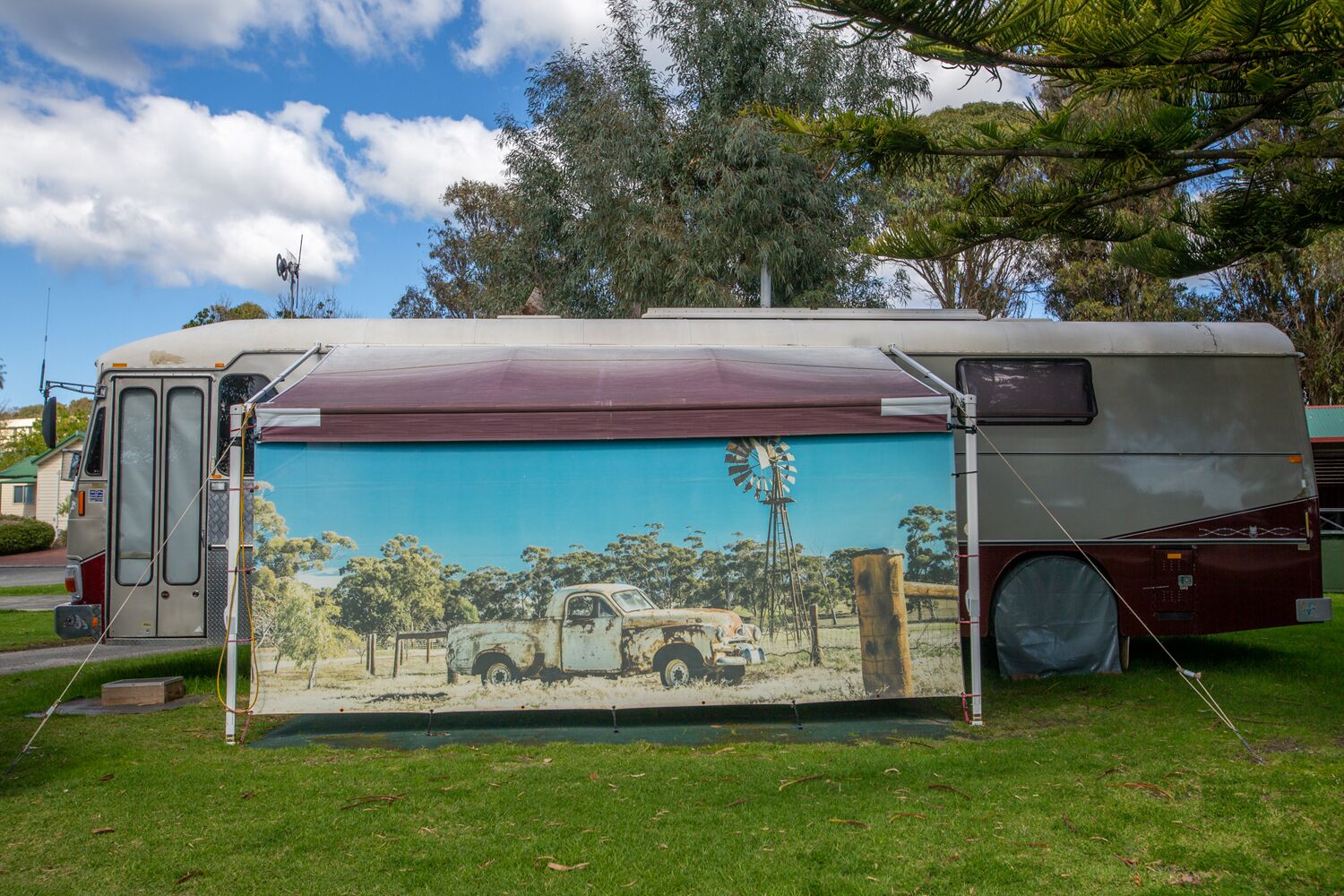 A vintage camper van parked on a grassy area with a mural of a rural scene on its side.