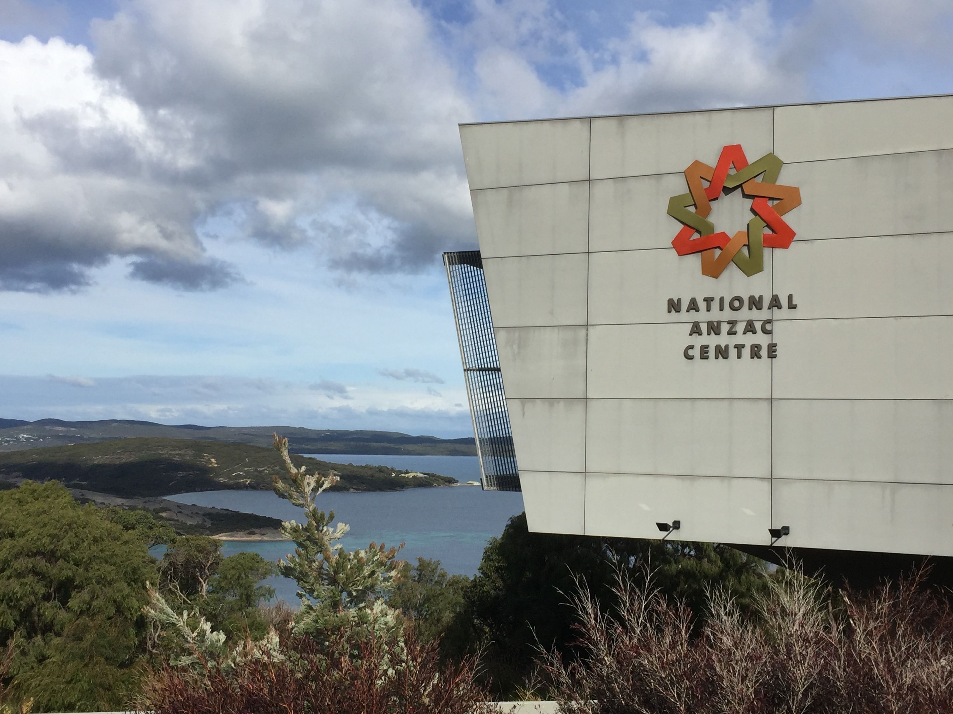 A sign for the National Anzac Centre with a scenic view of hills and a lake in the background.