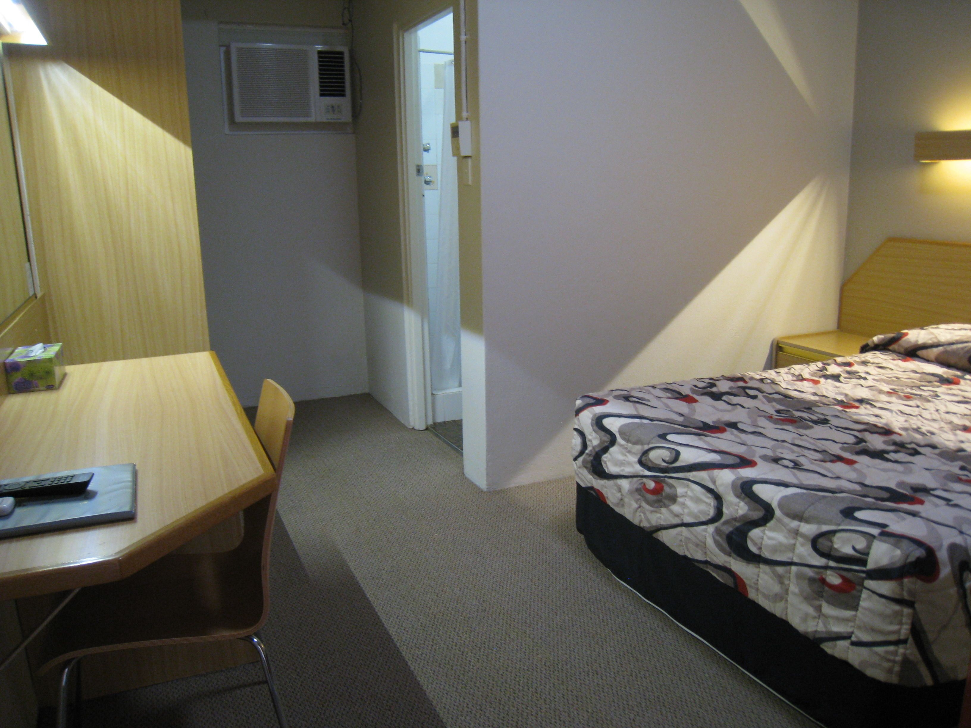 A hotel room with a bed, a desk, a chair, a bedside table, and a lamp.