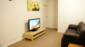Een woonkamer met een televisie op een houten standaard, een donkere leren bank en een schilderij van bloemen aan de muur.