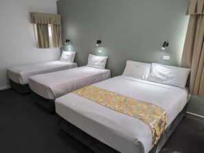 A hotel room with two single beds and one double bed, all with white bedding. There are wall-mounted lights and a window with curtains.