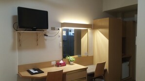 Een kleine kamer met een houten bureau, een stoel, een aan de muur bevestigde televisie, een spiegel en een kleine koelkast.