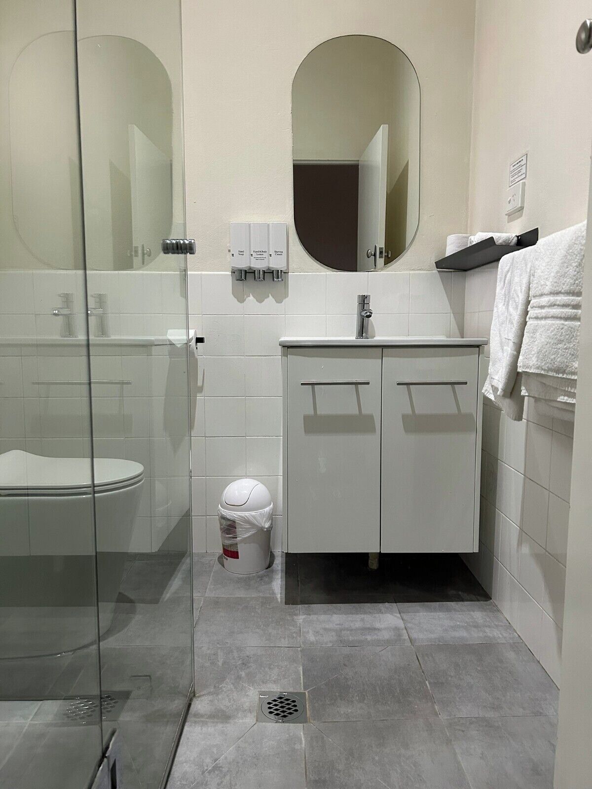 A modern bathroom with a glass shower enclosure, a white toilet, a sink with a mirror above, and hand soap dispensers.