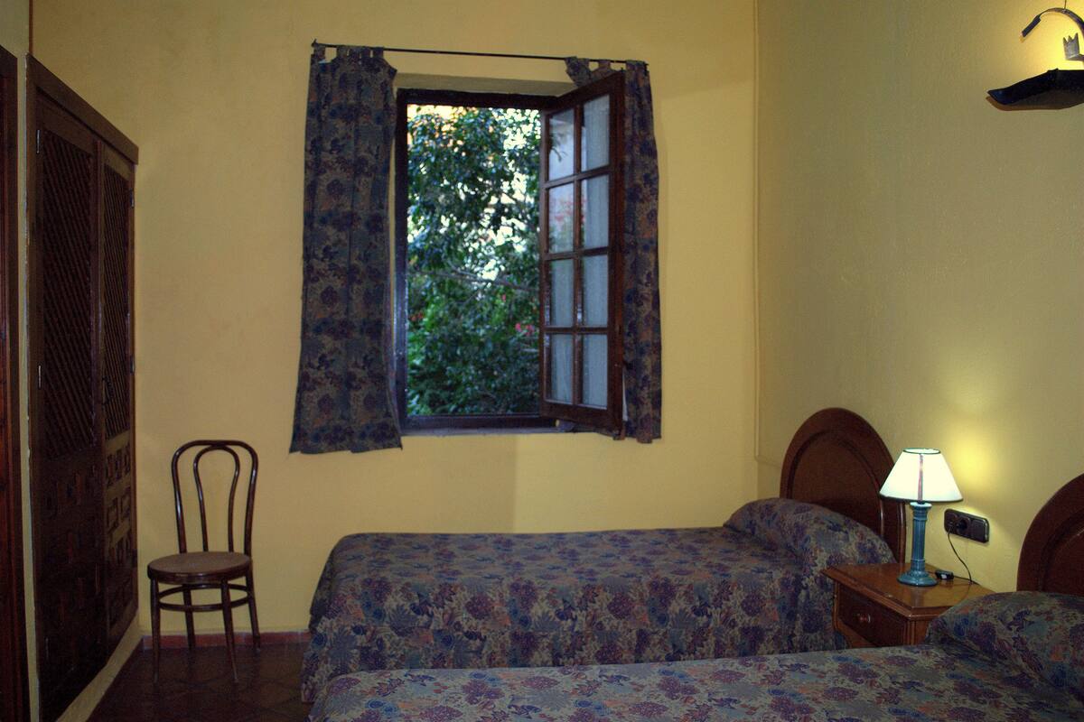 Un dormitorio con dos camas, una ventana con cortinas, una silla y una mesita de noche con lámpara.
