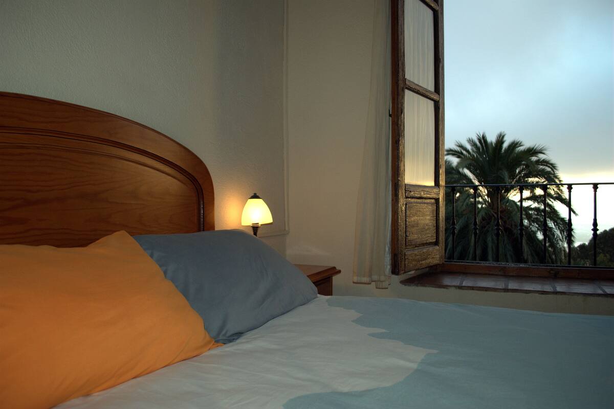 Un dormitorio con cabecera de madera, cama con ropa de cama blanca y azul, mesita de noche con lámpara y ventana con vistas a palmeras.