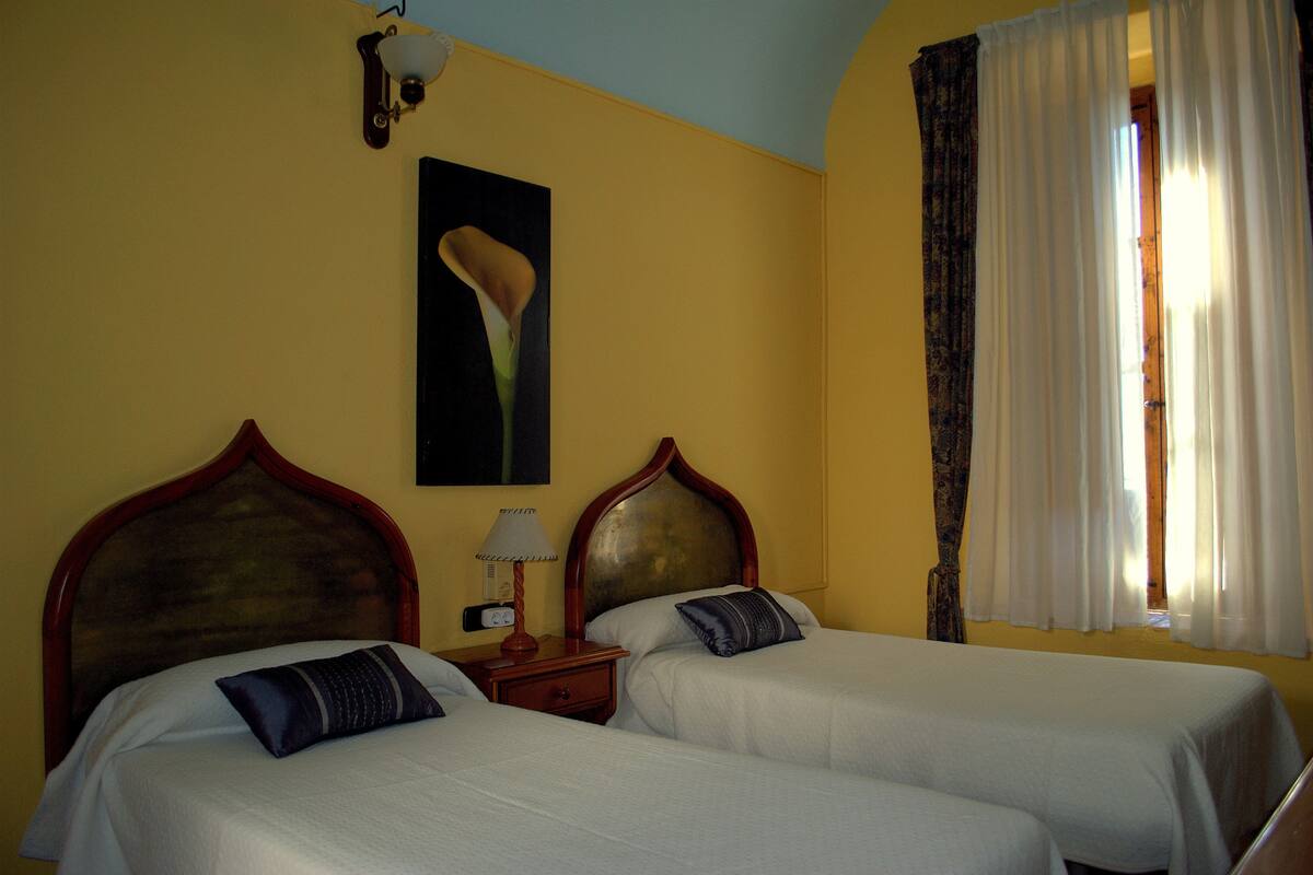 Habitación de hotel con dos camas, una mesita de noche con lámpara, un cuadro en la pared y una ventana con cortinas.