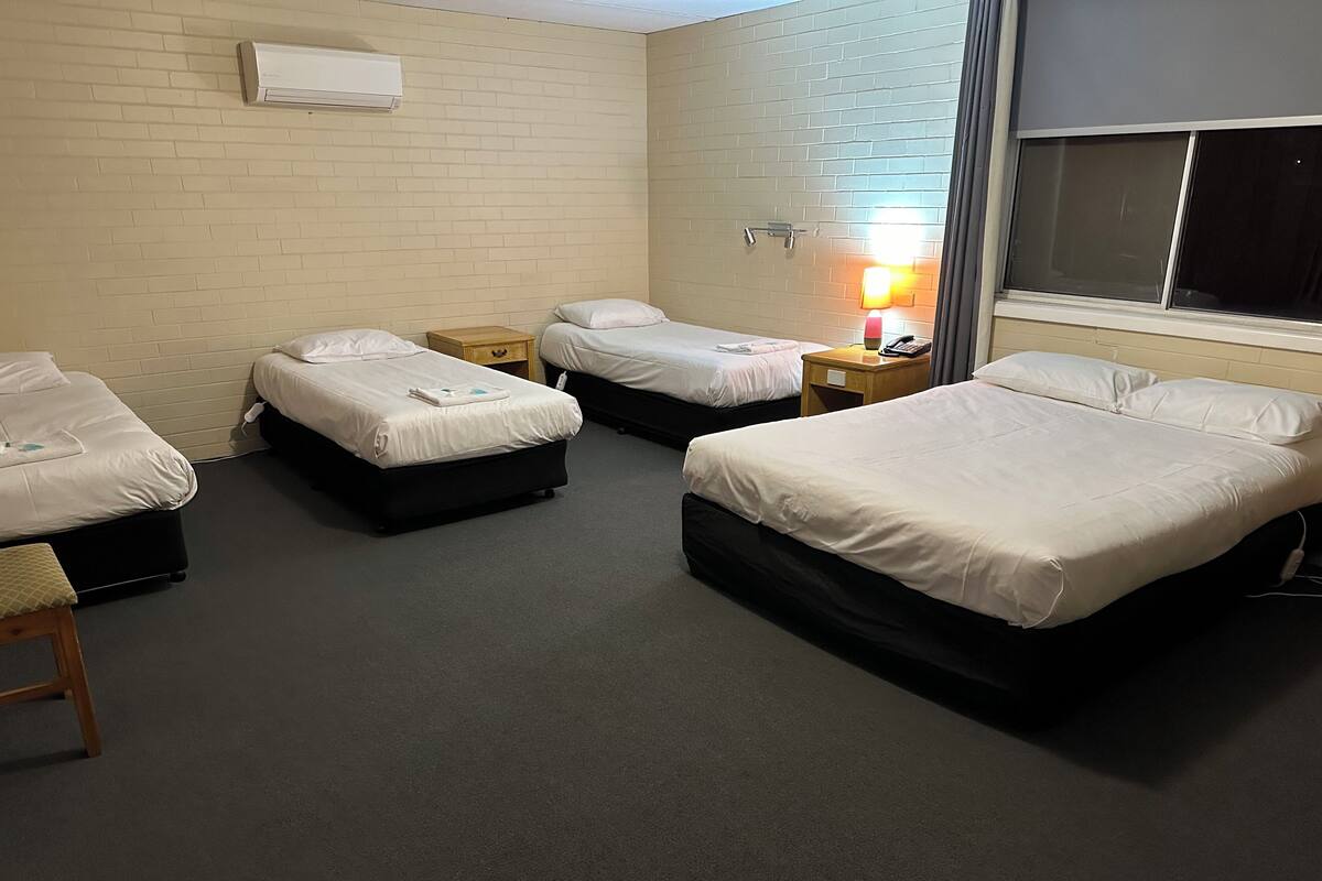 A hotel room with four beds, a wooden nightstand, a lamp, and a window with curtains.