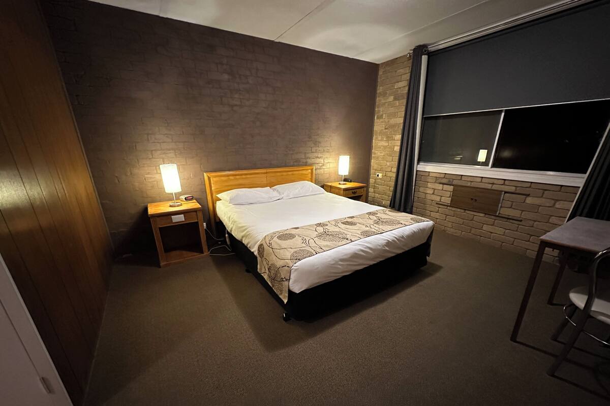 A hotel room with a single bed, two bedside tables with lamps, a window with curtains, and a small stool.