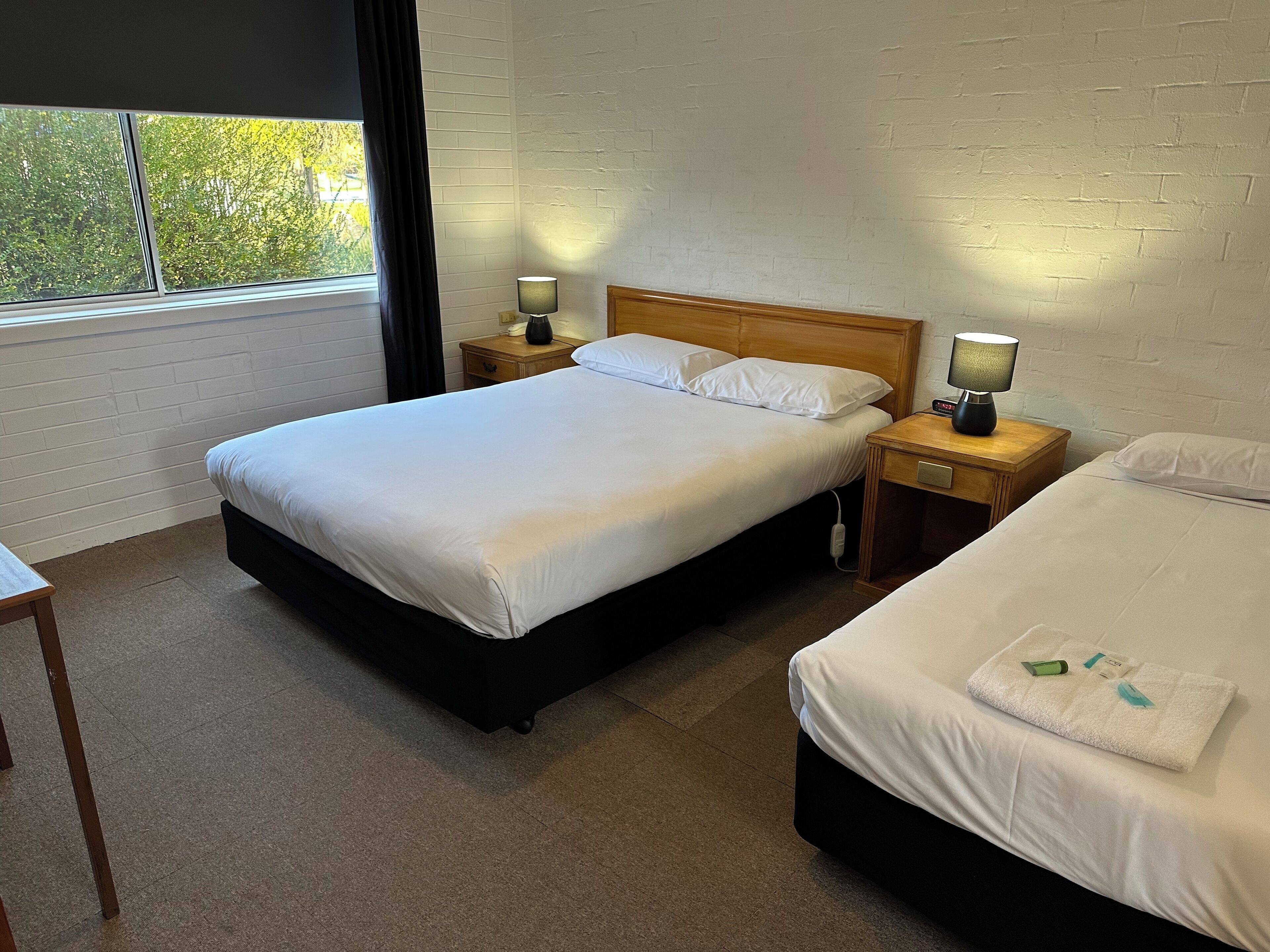 Une chambre d’hôtel avec un grand lit, un lit plus petit, deux tables de chevet avec des lampes, une fenêtre avec des rideaux et un mur en briques blanches.