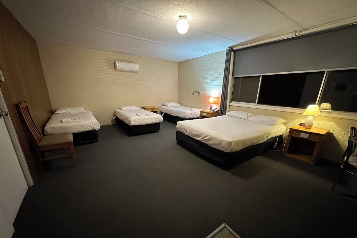 A hotel room with four single beds, a wooden chair, a desk with a lamp, a window, and a wall-mounted air conditioner.