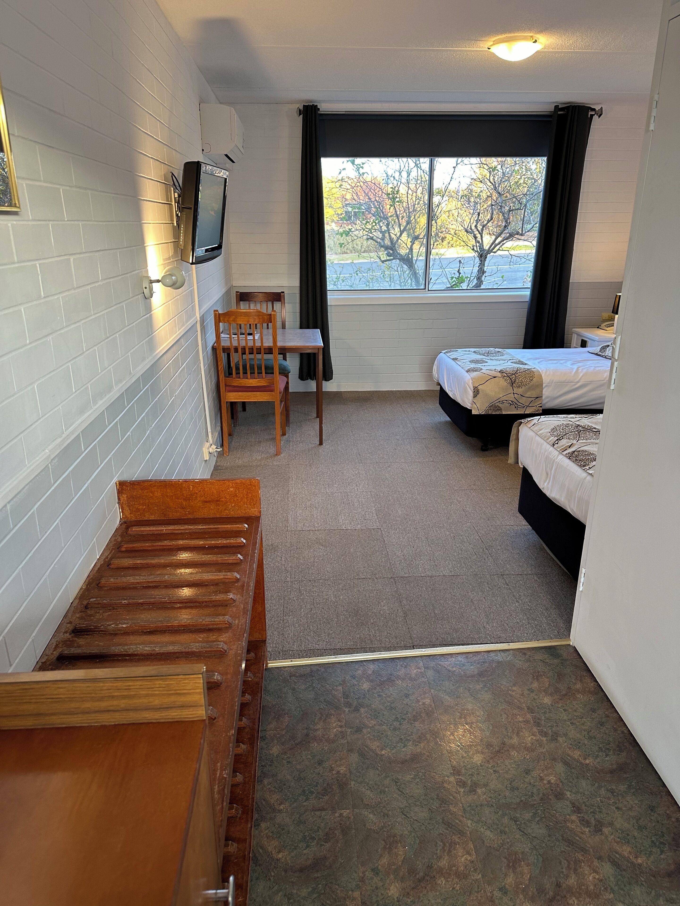 A small, compact room with a bed, a desk, and a chair. There is a window with a view of trees and a building outside.
