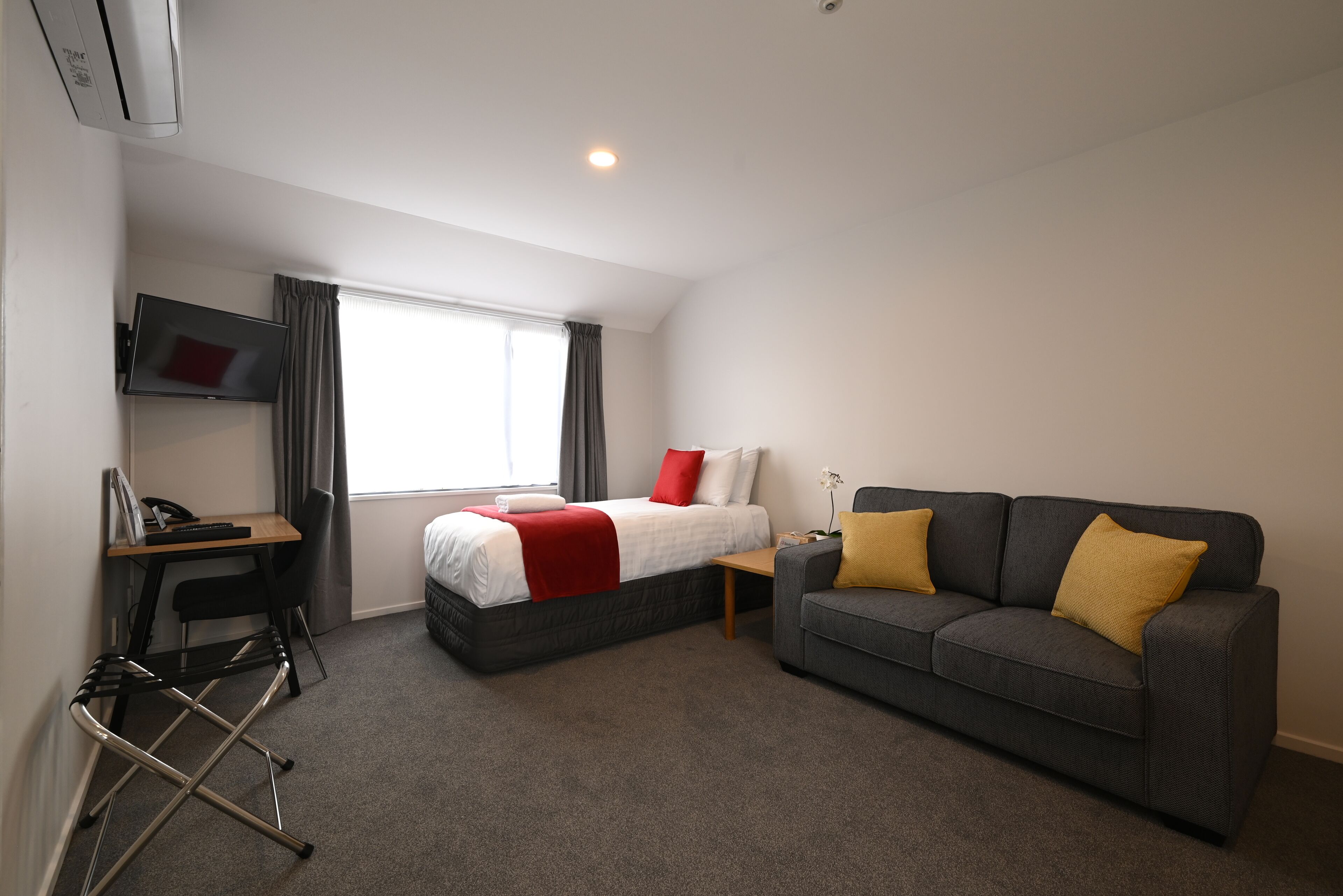 Quarto de hotel com uma cama, um sofá cinzento, uma escrivaninha com uma cadeira e uma televisão na parede.