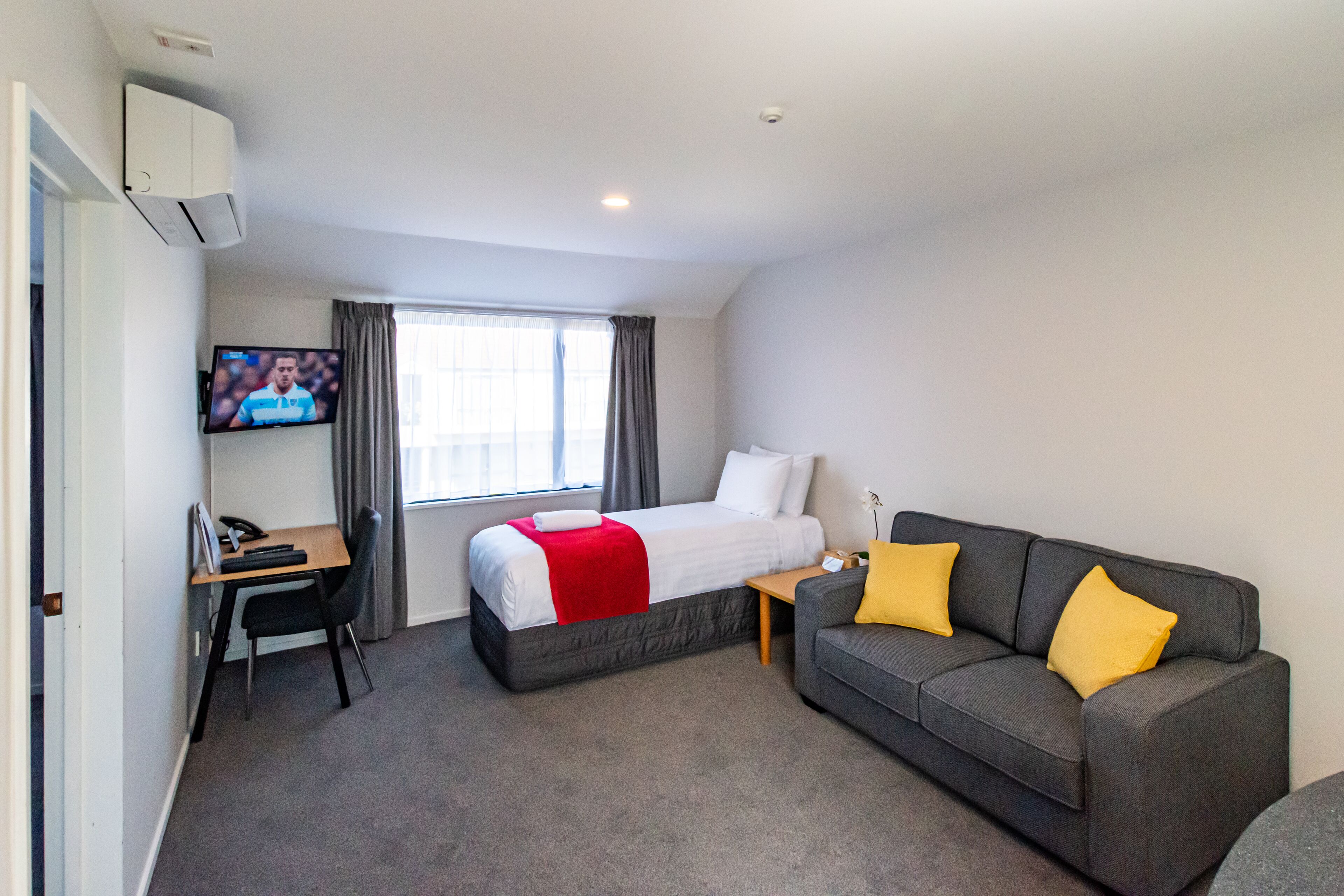 Une chambre d’hôtel avec un lit, un bureau, un canapé gris et un téléviseur fixé au mur.