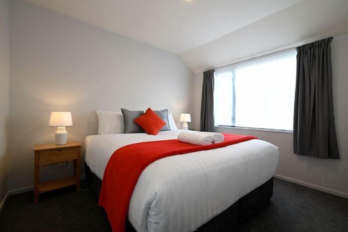 Ein Hotelzimmer mit einem großen Bett, zwei Nachttischen, einem fenster mit Vorhängen und einer Lampe auf jedem Tisch.