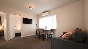 A compact living space with a sofa, a dining table with chairs, a TV mounted on the wall, and an air conditioning unit. - Riccarton Mall Motel (Christchurch)