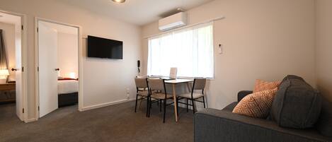 Un espacio habitable compacto con sofá, una mesa de comedor con sillas, un televisor montado en la pared y un equipo de aire acondicionado.