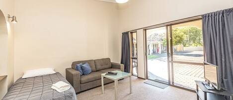 A room with a bed, a sofa, a small table, a TV on a stand, and a sliding glass door leading outside.