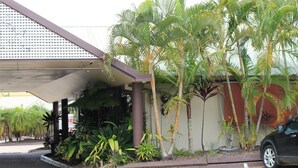 A hotel entrance with a covered parking area, palm trees, and a black car parked nearby. - Glenmore Palms Motel (Norman Gardens)