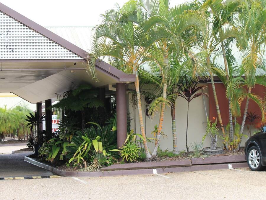 Entrada de un hotel con zona de estacionamiento techada, palmeras y un coche negro aparcado cerca.