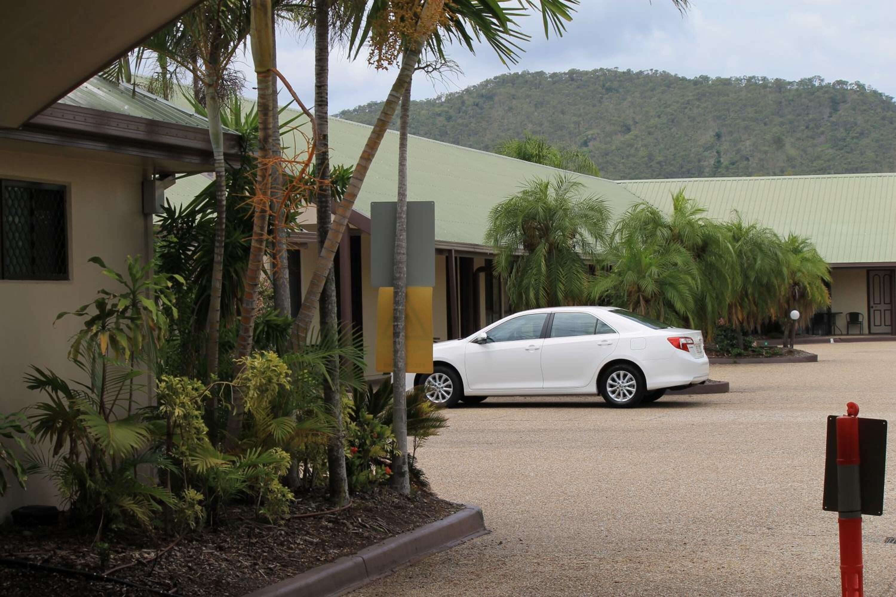 Un edificio con un coche estacionado frente a él, rodeado de plantas tropicales y un paisaje montañoso al fondo.