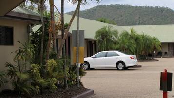Un edificio con un coche estacionado frente a él, rodeado de plantas tropicales y un paisaje montañoso al fondo.