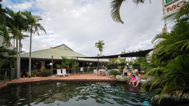 A pool area with lounge chairs and palm trees, surrounded by a covered patio and a building with a green roof. - Glenmore Palms Motel (Norman Gardens)