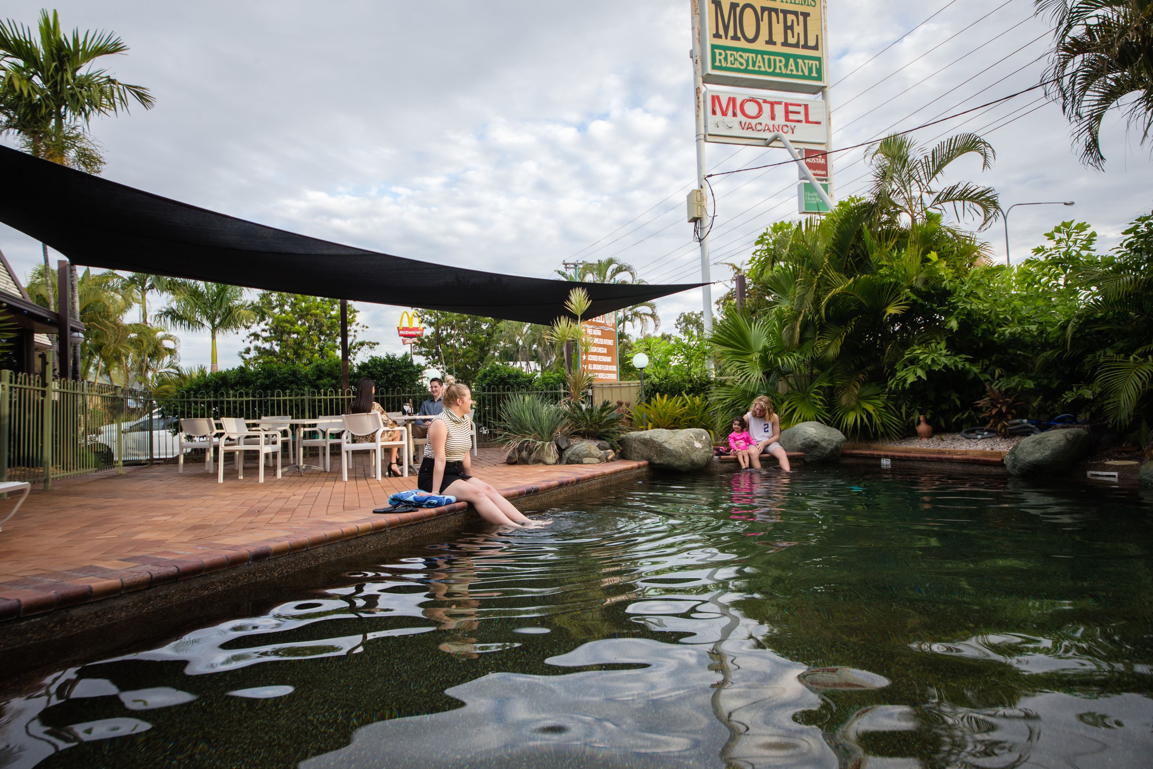 A swimming pool area with a shaded patio, palm trees, and a motel sign.