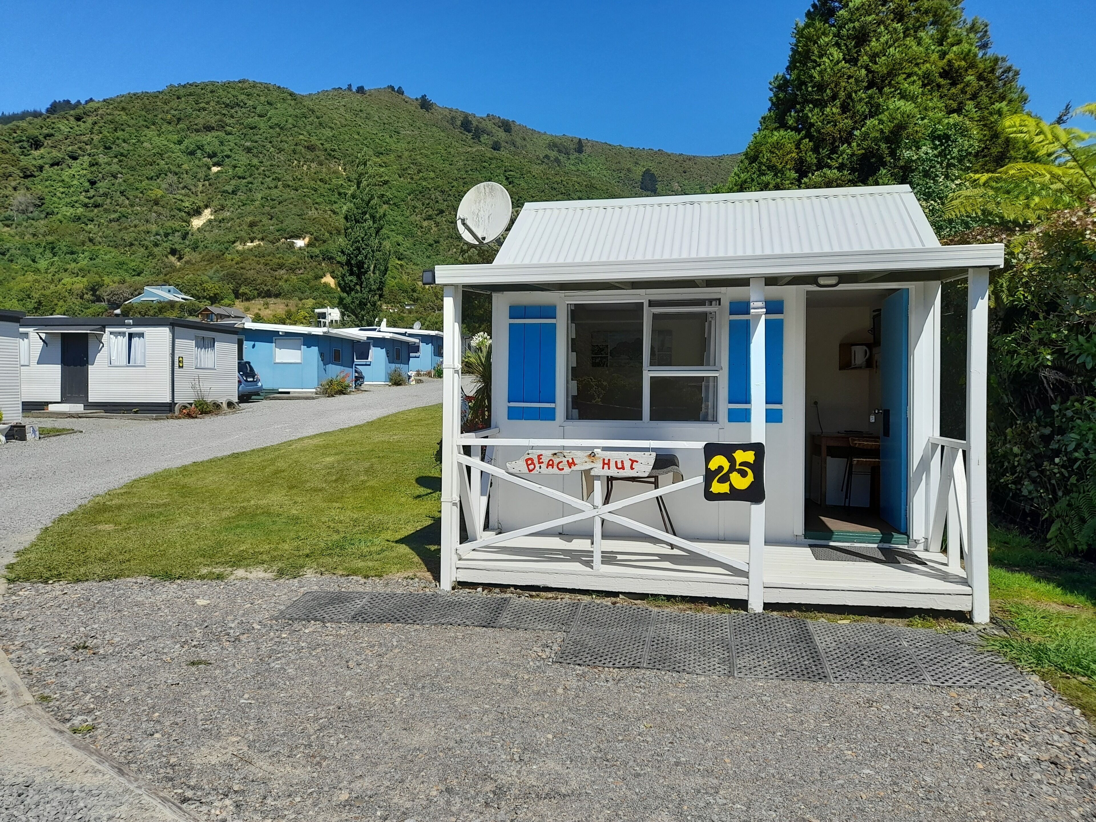 A small white building with blue shutters, labeled "BEACH HUT," located in a campground with multiple blue and white cabins in the background.