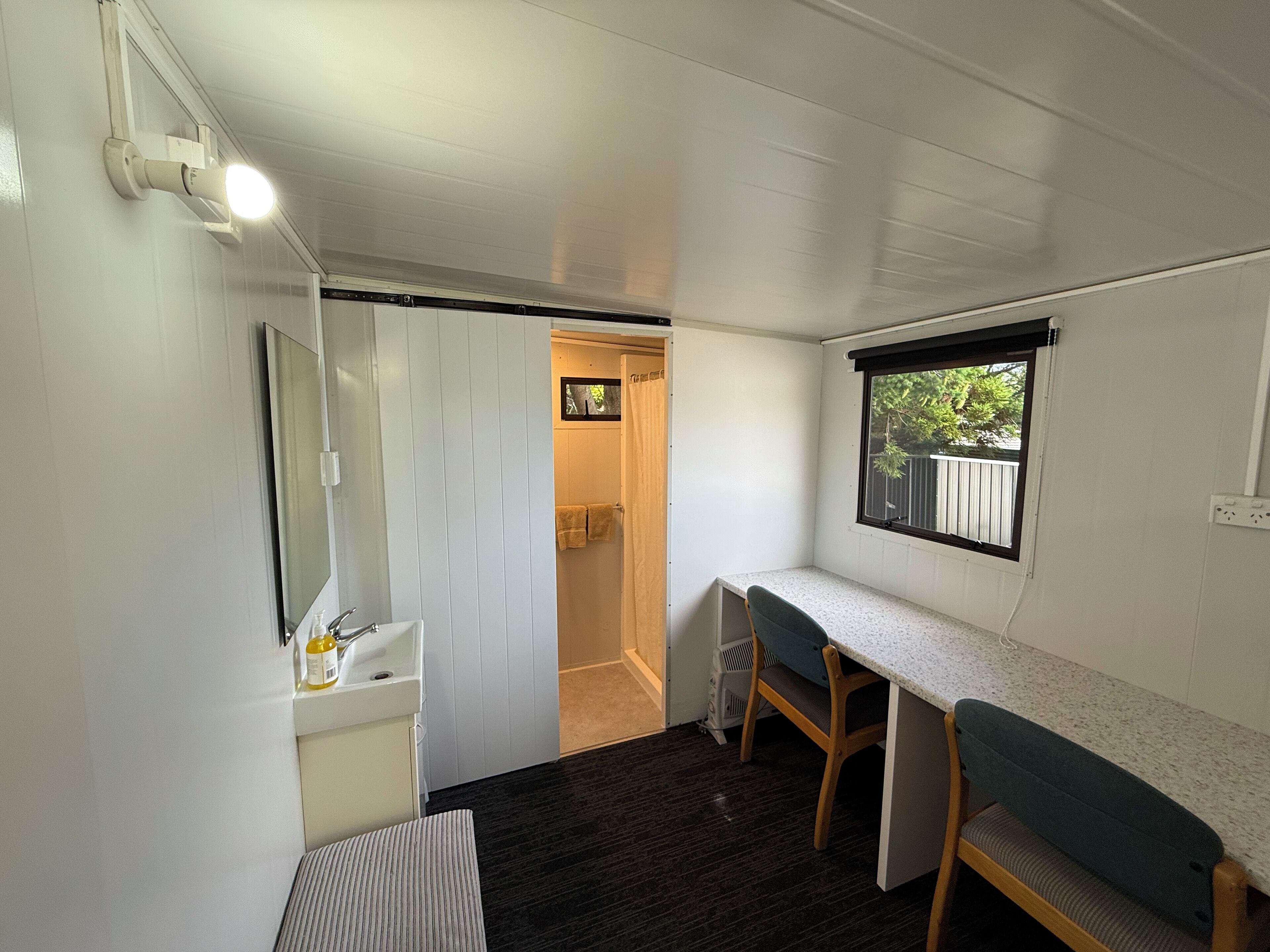A compact, well-lit room with a kitchenette, a small seating area, and a window providing a view of the outdoors.