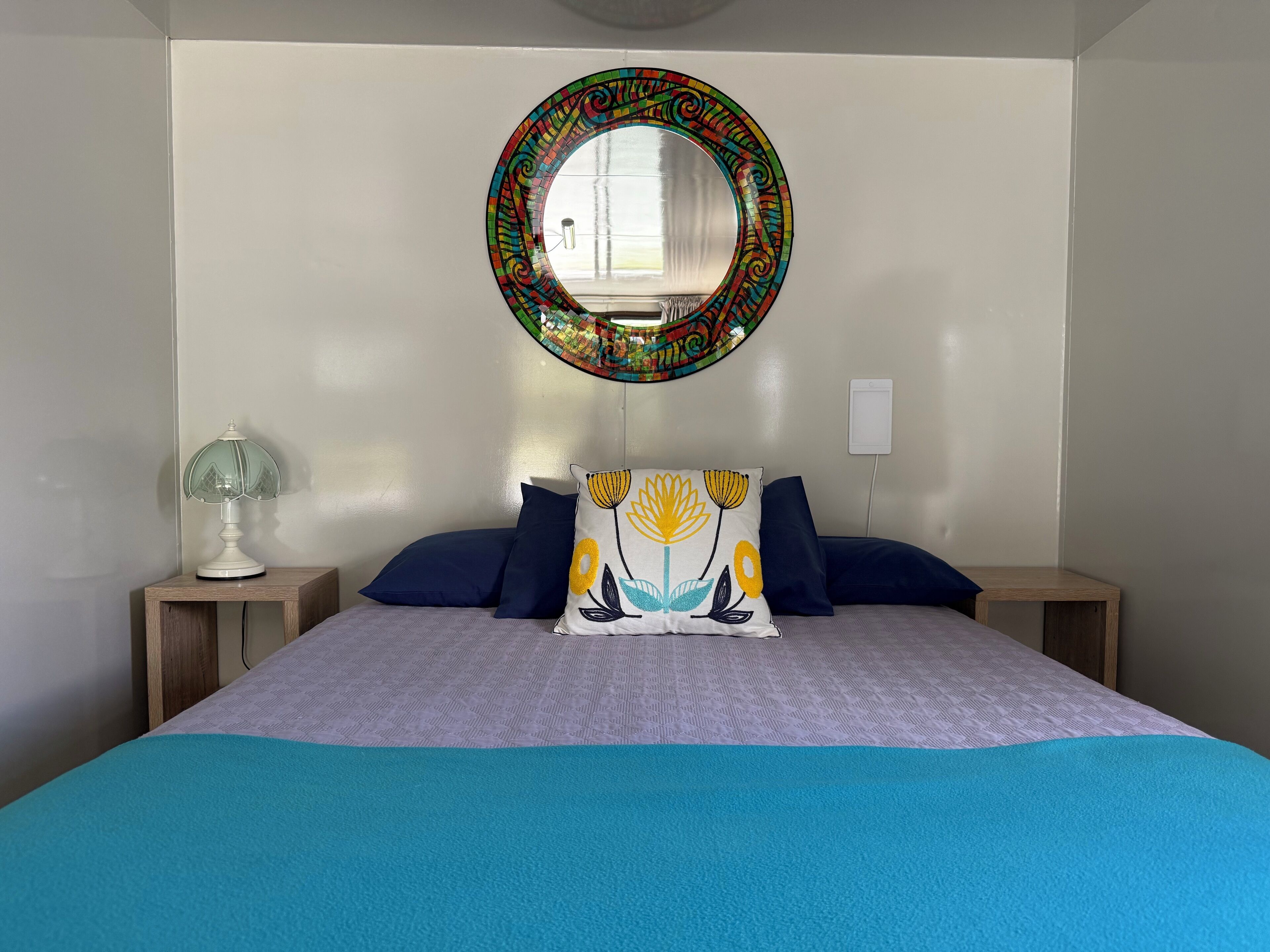 A bed with a blue blanket, two pillows, a bedside lamp, and a wall-mounted mirror with a colorful frame.