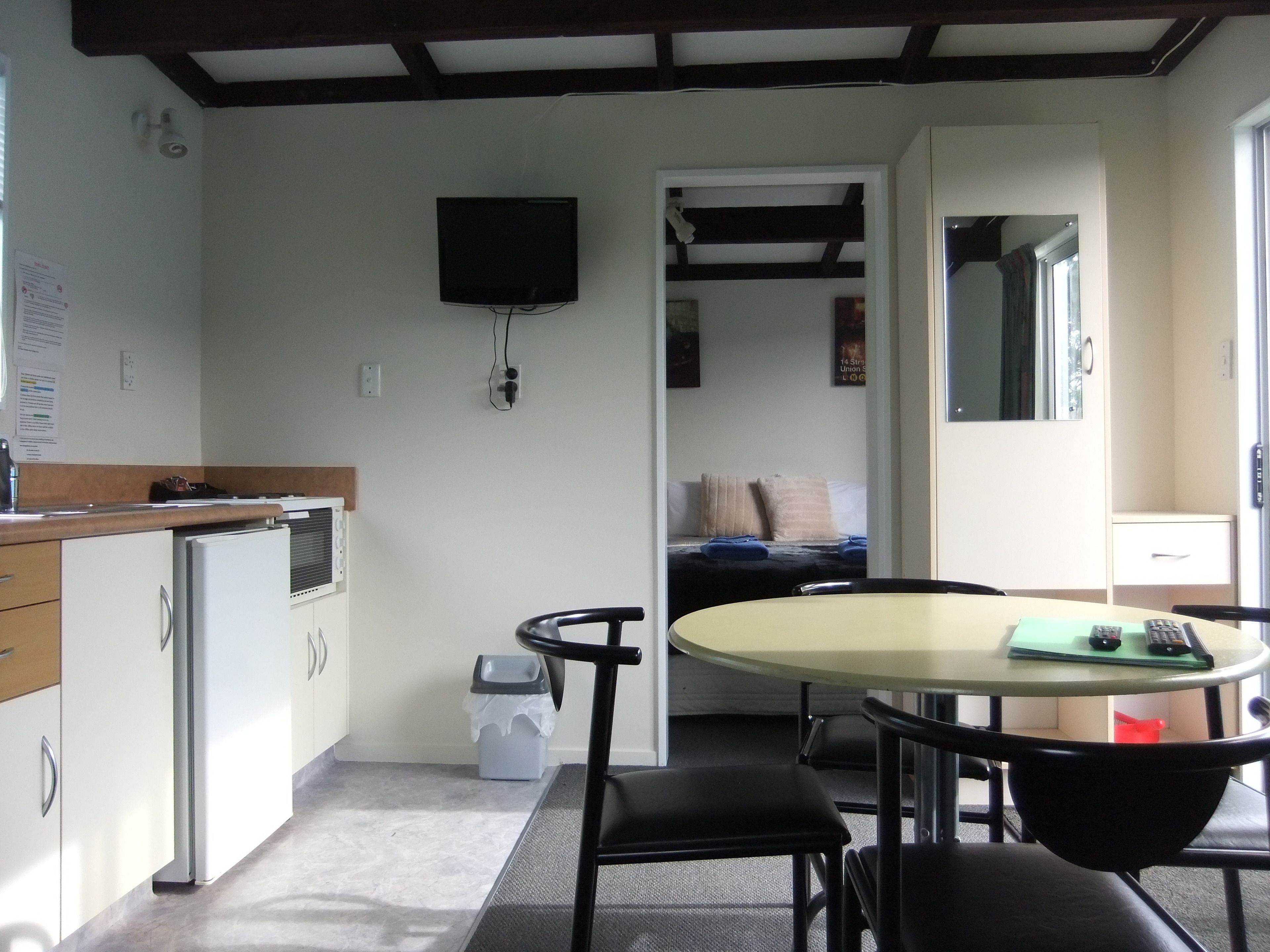 A compact kitchen with a refrigerator, a small sink, and a microwave. There is a dining area with a round table and chairs. A television is mounted on the wall.