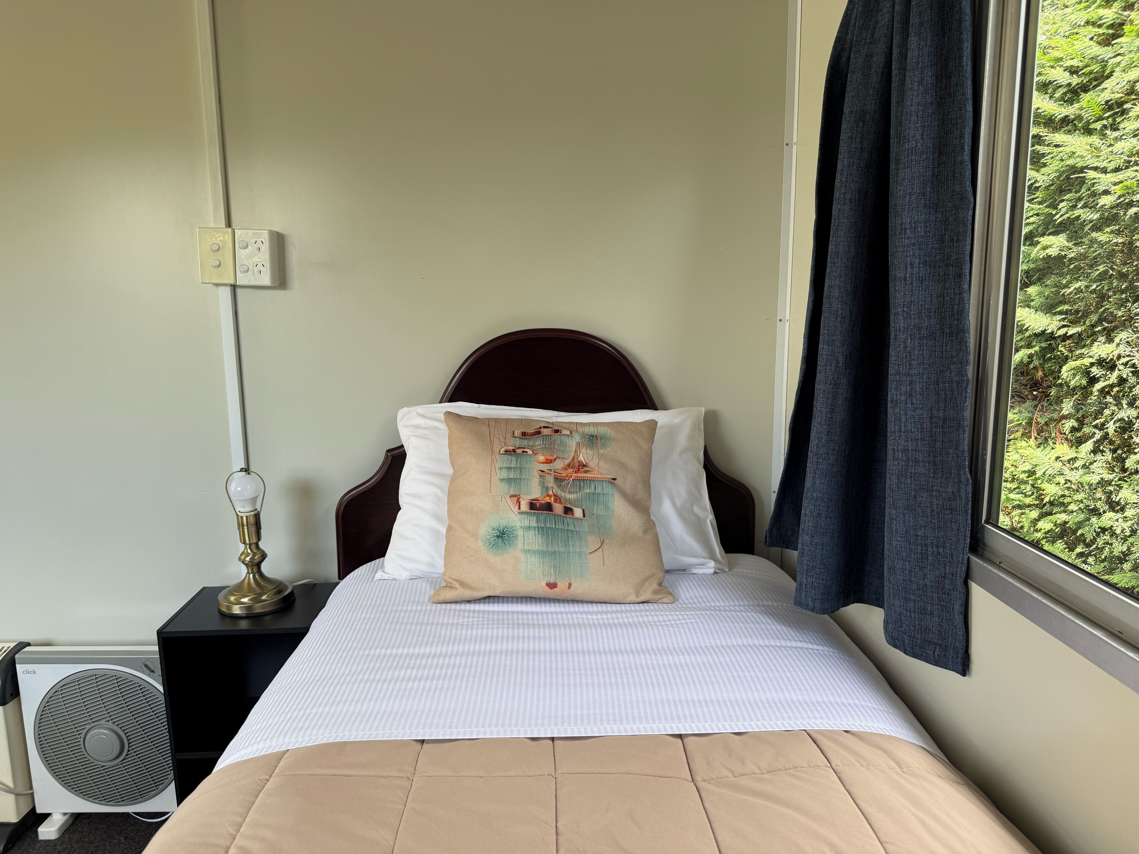 Un letto con un cuscino bianco e un cuscino decorativo, un comodino con una lampada, una ventola e una finestra con tende.