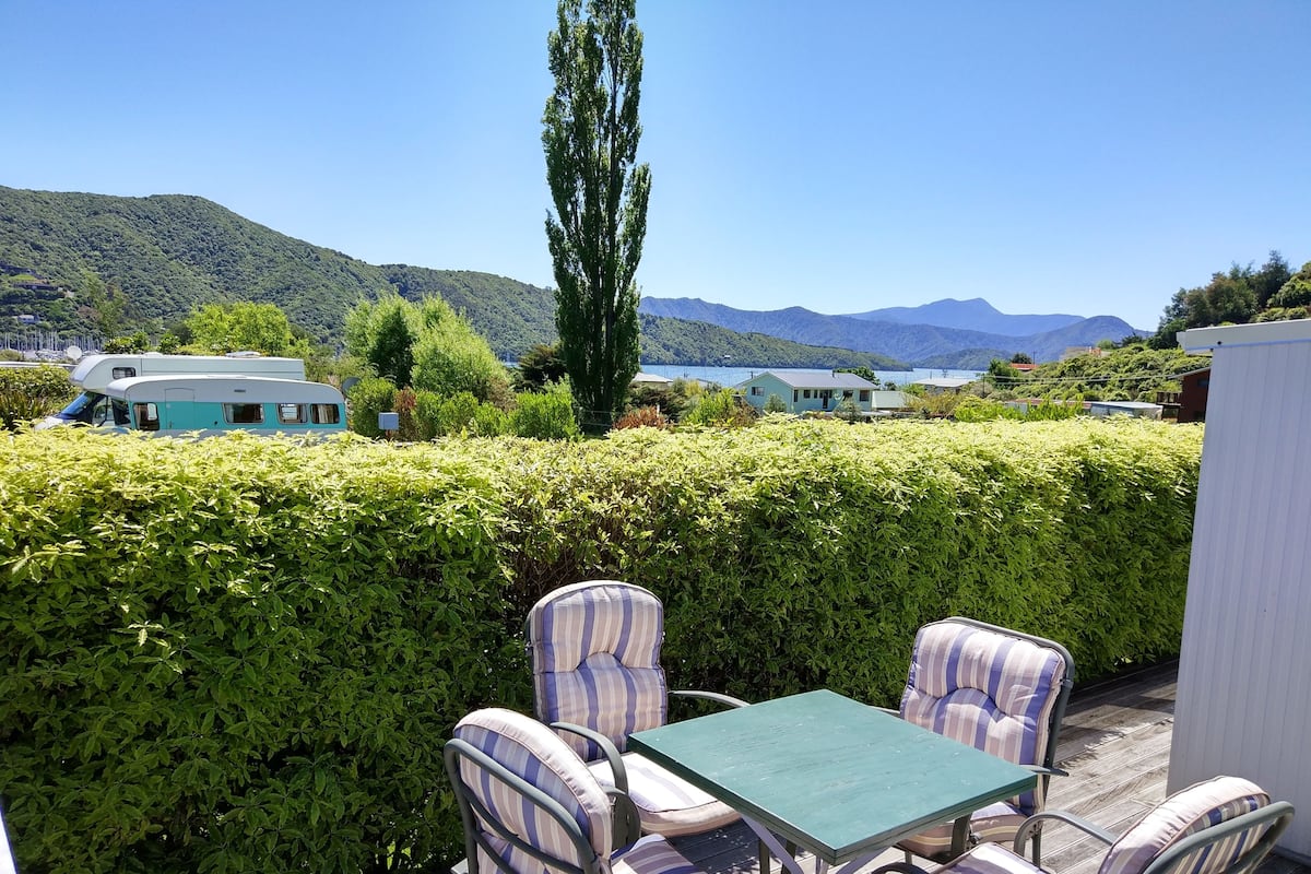 An outdoor patio area with striped cushioned chairs and a table, surrounded by a hedge and overlooking a scenic landscape with mountains and a body of water.