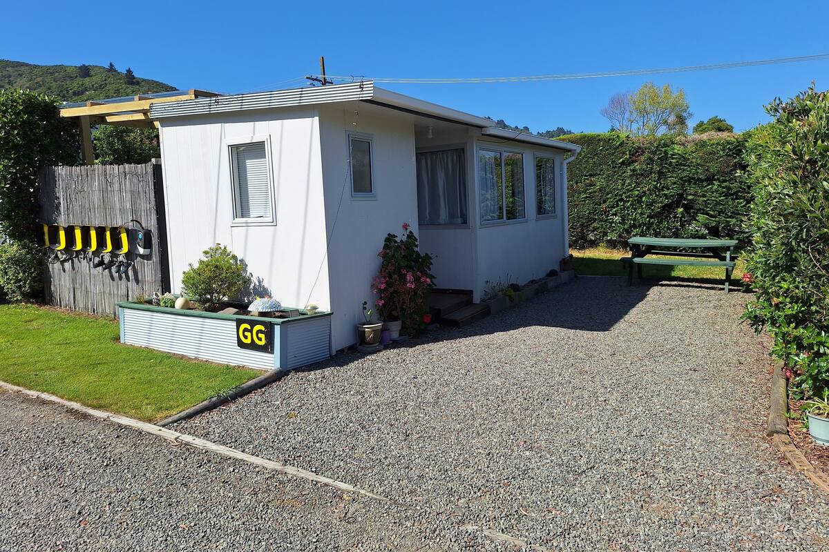 A small, single-story house with a gravel driveway, a wooden fence, and a green picnic table.