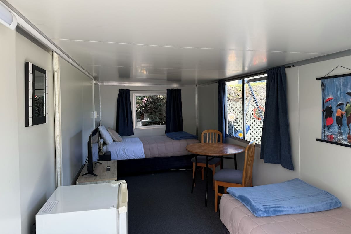 A compact room with a bed, a small table, and a chair. There is a window with curtains, a wall-mounted mirror, and a framed picture.