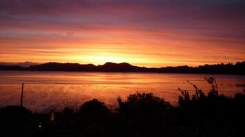 Un atardecer sobre un lago tranquilo, con siluetas de árboles y una colina distante.