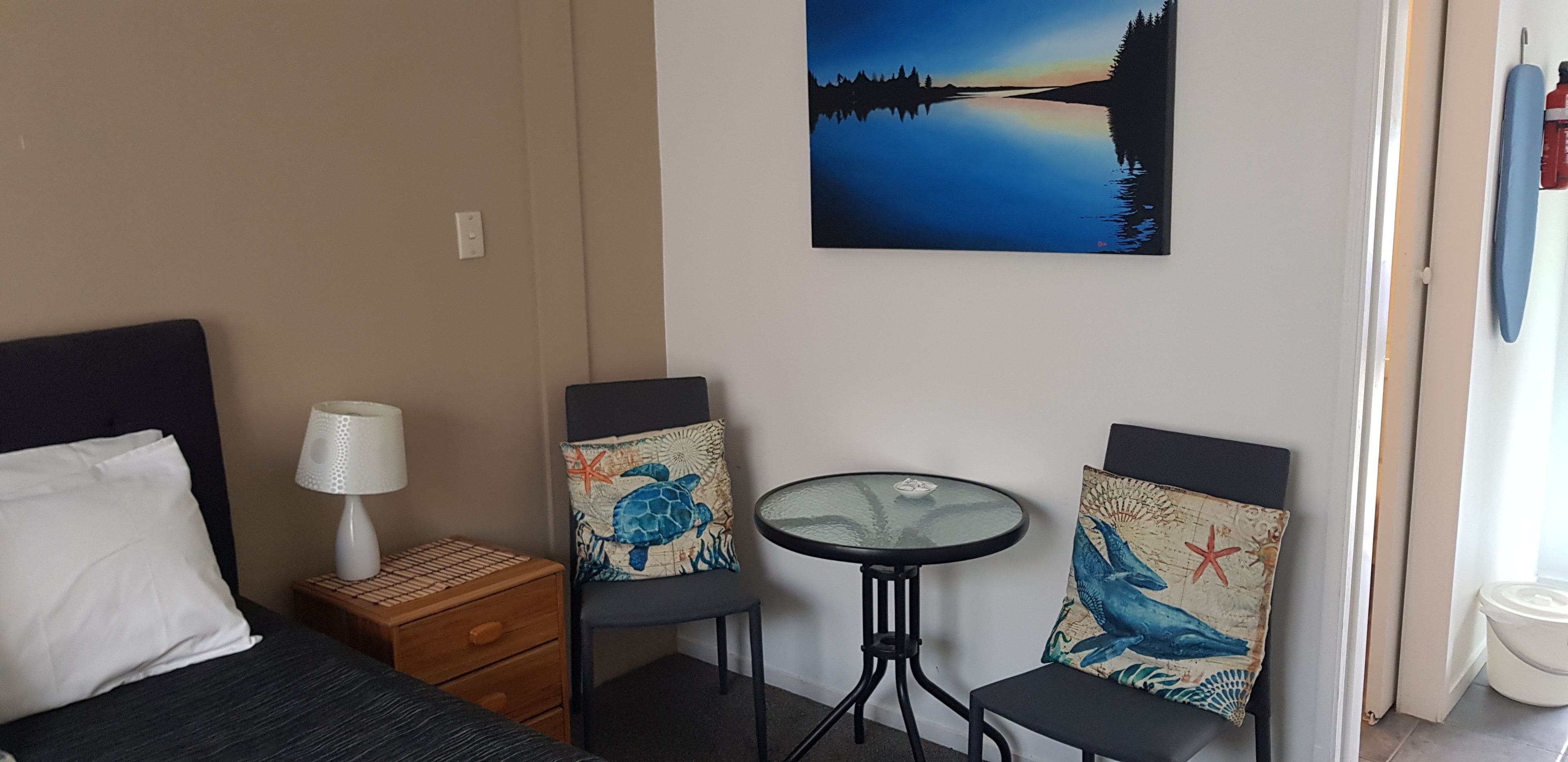 A hotel room with a bed, a nightstand with a lamp, two chairs with decorative pillows, a small round table, and a painting of a lake scene on the wall.