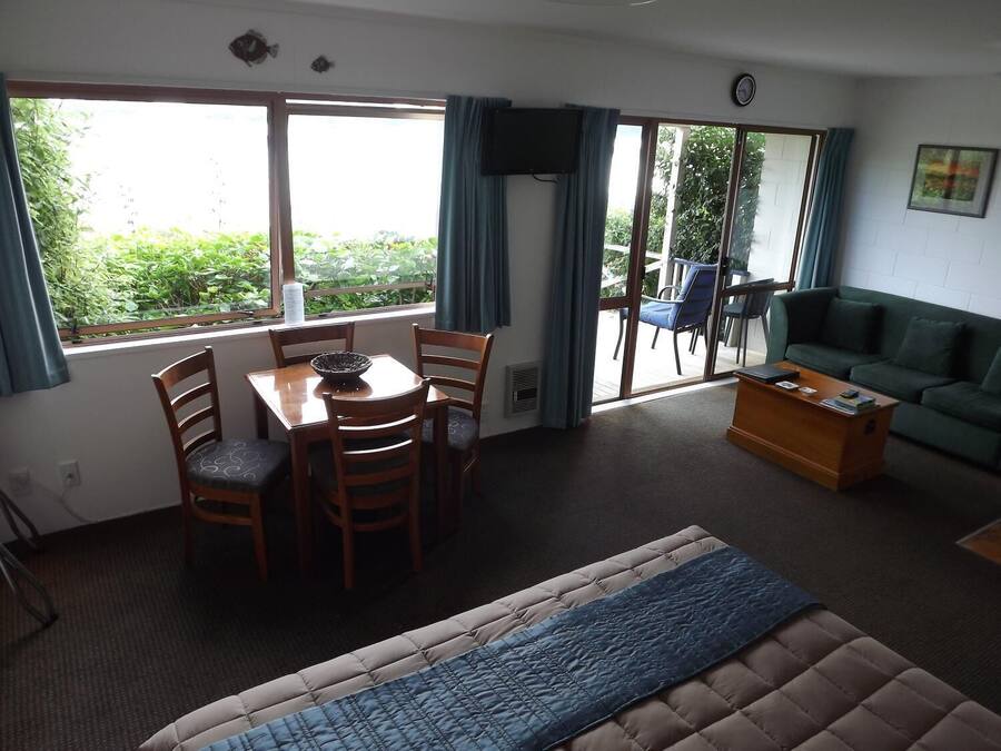 Una habitación con una mesa de comedor, sillas, un sofá, una mesa de centro y un televisor. Hay ventanas con cortinas y vistas al exterior con áreas verdes.